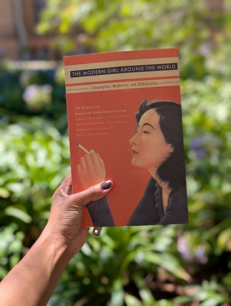 A hand holding a book in the leafy area that appears in the background. The Modern Girl Around the World book. 